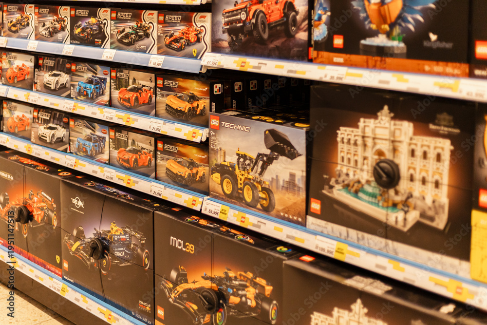 Naklejka premium Chemnitz, Germany - March 14, 2026: Shelves in a retail store show many Lego building kits for sale. The kits include cars, buildings, and construction vehicles. Shoppers browse the toy section