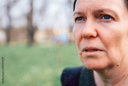 Close-up portrait of mature woman with blue eyes looking thoughtfully off-camera, natural light and blurred green park background