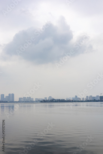 Calm lake with Hanoi skyline in the distance under overcast sky showing urban waterfront, peaceful atmosphere, and minimal cityscape composition with reflections and soft tones