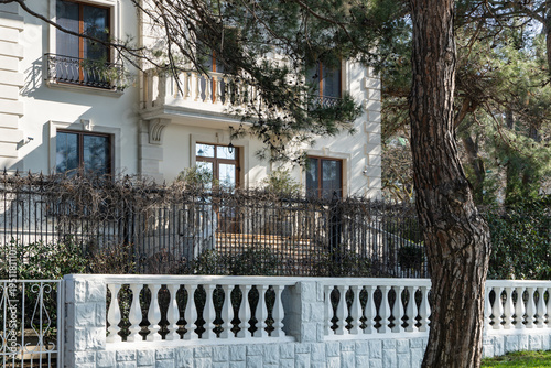 Stylish two-story building with balcony, framed by tall trees and  decorative metal fence, featuring elegant white stone balusters and large windows. Gelendzhik Bay, Black Sea. March 2026