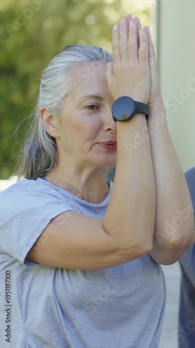 Vertical video: Woman following cue, raising and lowering palms on porch, wearing gray tee, watch