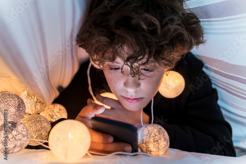 Boy with chain of lights underneath bedcover using cell phone