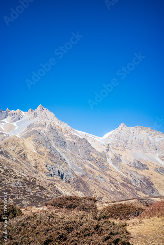 Clean and minimalist composition of rugged Himalayan mountain peaks rising above dry alpine vegetation under a vast, cloudless blue sky. The image highlights dramatic ridgelines, rocky textures