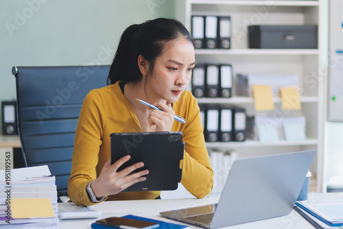 Asian woman works on laptop at office desk. She smiles happily while typing on keyboard near window with blinds. Professional woman works in business environment.