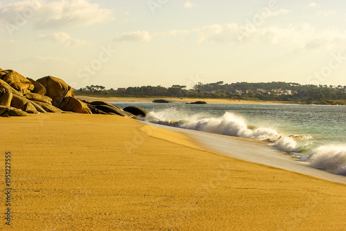 Tableau sur toile Golden sand beach with waves crashing ashore and a village nestled in the trees on the hill in the background