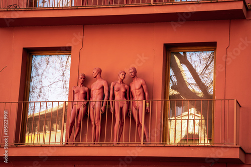Four red mannequins on a red balcony in front of a red facade with windows and reflections.
