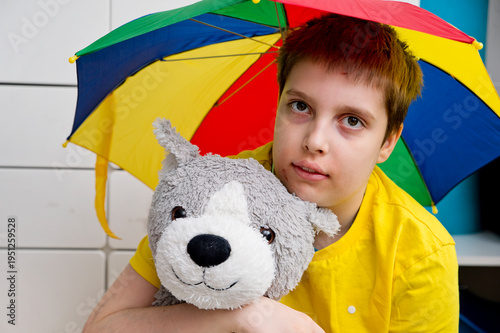 An autistic boy holds a colorful umbrella and a large plush toy. April is Autism Awareness Month.