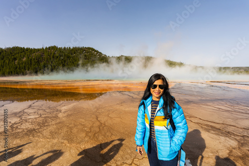 A portrait of a smiling woman in a blue quilted jacket and sunglasses standing at the edge of the Grand Prismatic Spring.