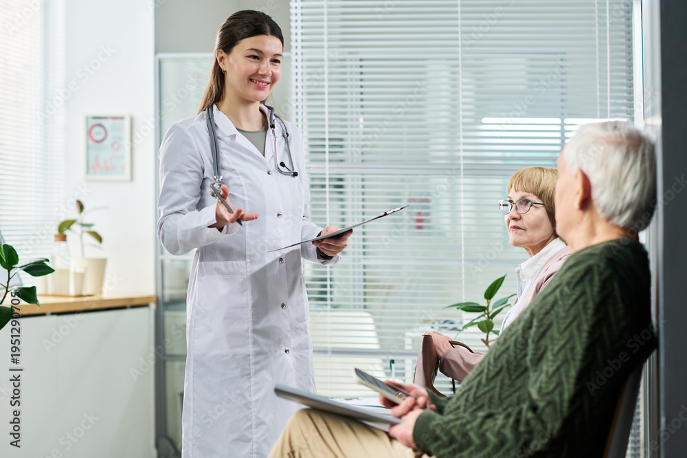 Obraz premium Young female doctor smiling while talking to senior man and senior woman sitting in medical office waiting area, holding clipboard and digital tablet