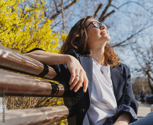 Relaxed young curly woman in glasses enjoying spring sun on a city bench under blooming yellow forsythia bushes