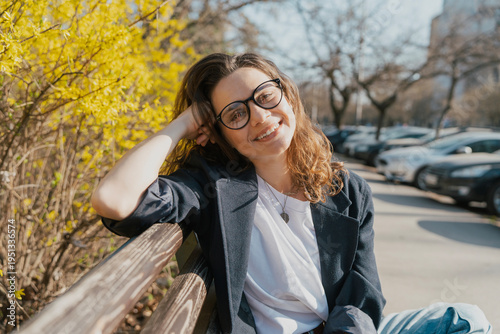 Relaxed young curly woman in glasses enjoying spring sun on a city bench under blooming yellow forsythia bushes
