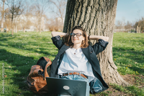 Relaxed young woman sitting on green grass in a city park with laptop and enjoying the sun.