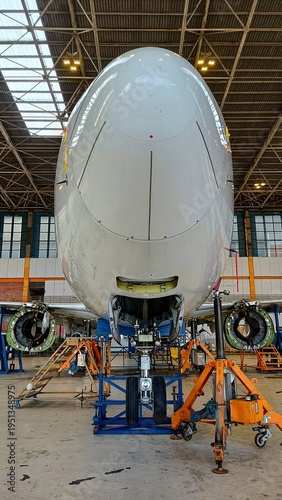 Front view of a commercial airplane undergoing maintenance inside a hangar, with engines removed and nose landing gear supported by equipment, showcasing aviation engineering and repair process