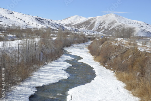 a winding river flowing through a snowy valley under a clear blue sky the water is partially bordered by thick white ice and snow 