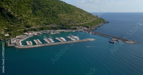 Aerial view of a scenic coastal harbor at the base of a lush, green mountain. Numerous boats are docked within the calm marina, where the deep blue sea meets the verdant, tree-covered slopes.
