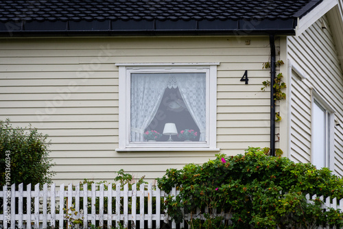 View of a cozy window adorned with lace curtains, a lamp, and flowers, nestled in a quaint house with a white picket fence, Lofoten Islands, Nordland, Norway.