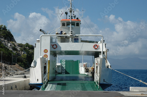 A white passenger and car ferry with an open ramp docked at a stone pier in a Mediterranean harbor under a blue sky