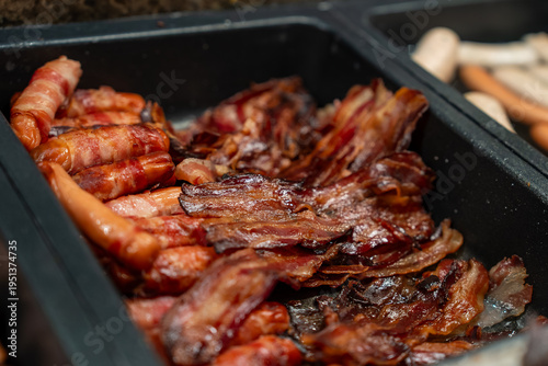 Trays hold bacon strips and bacon wrapped sausages in a buffet setup. Warm directional lighting highlights caramelized edges and rich textures in a luxury hotel setting.