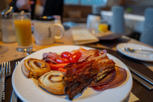 Crispy bacon, cured meats, cherry tomatoes, and two mini pastry swirls sit on a neat table with silverware, orange juice, and a branded mug in warm morning light.