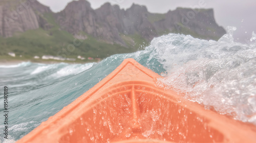 The bow of an orange kayak cuts through a wave by a mountain shore.