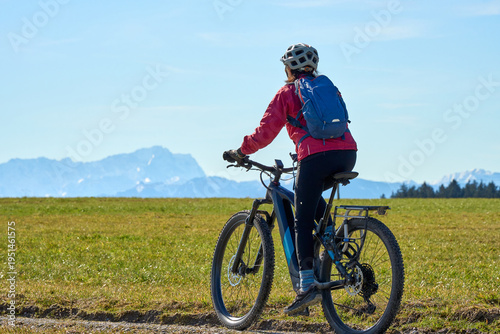 Middle aged woman cycling on an electric bike across green spring meadow in Upper Bavaria with alpine mountain range and Zugspitze silhouette in background under clear blue sky.