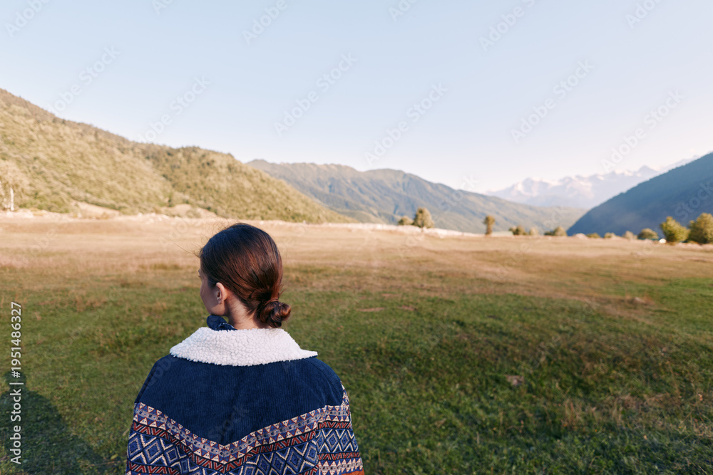 Fototapeta premium Woman back portrait in sweater standing in open field, looking at mountain landscape and distant horizon. Nature scene with valley, green grass and clear sky for travel and solitude.