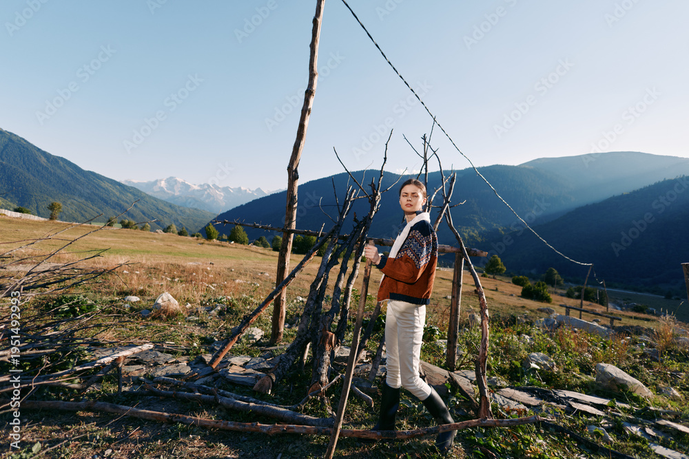 Fototapeta premium Woman in sweater and boots standing by a rustic fence in a rural field with mountains in the background, countryside landscape and clear sky for outdoor lifestyle travel