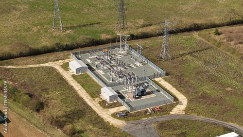 Aerial view of an electrical substation standing stark against the green landscape, power lines reaching into the bright sky, Retford, United Kingdom.