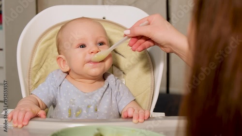 Caring mother feeding her cute infant son his first solid food from a spoon. The little toddler is sitting in a high chair, eating vegetable puree for lunch and getting his face and clothes messy