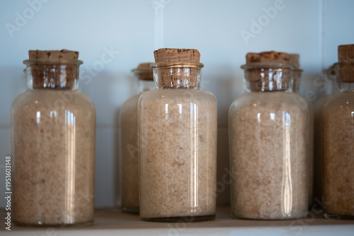 A row of glass bottles filled with salt and sealed with cork stoppers occupies the foreground against a subtly textured white background
