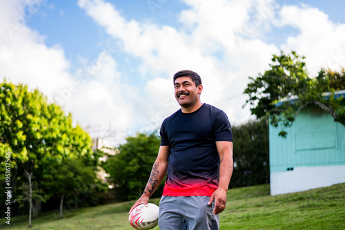 A young man walks cheerfully in a park, holding a rugby ball in the sunlight with trees around