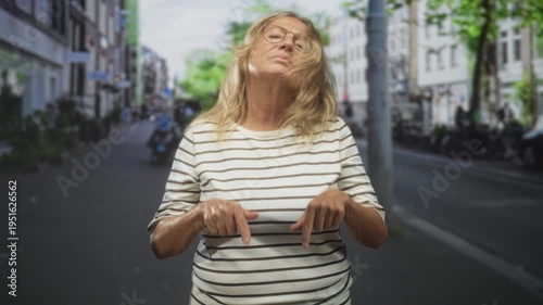 Senior woman pointing both index fingers down at pavement on a city street wearing striped shirt; quiet contemplation.
