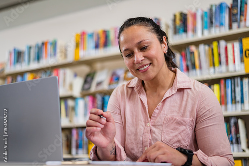 A woman is studying on a laptop in a library setting with bookshelves behind her