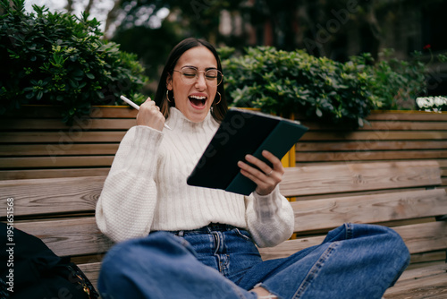 Overjoyed female designer rejoicing success while working on digital tablet outdoors, young woman in glasses and white sweater expressing triumph and happiness, startup victory and achievement.