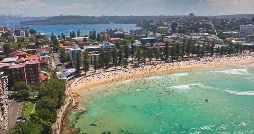 Crowds people on Manly Beach enjoying a sunny summer day, turquoise waters and sandy shore framed by coastal buildings and lush greenery in an aerial panoramic view. Travel to Australia