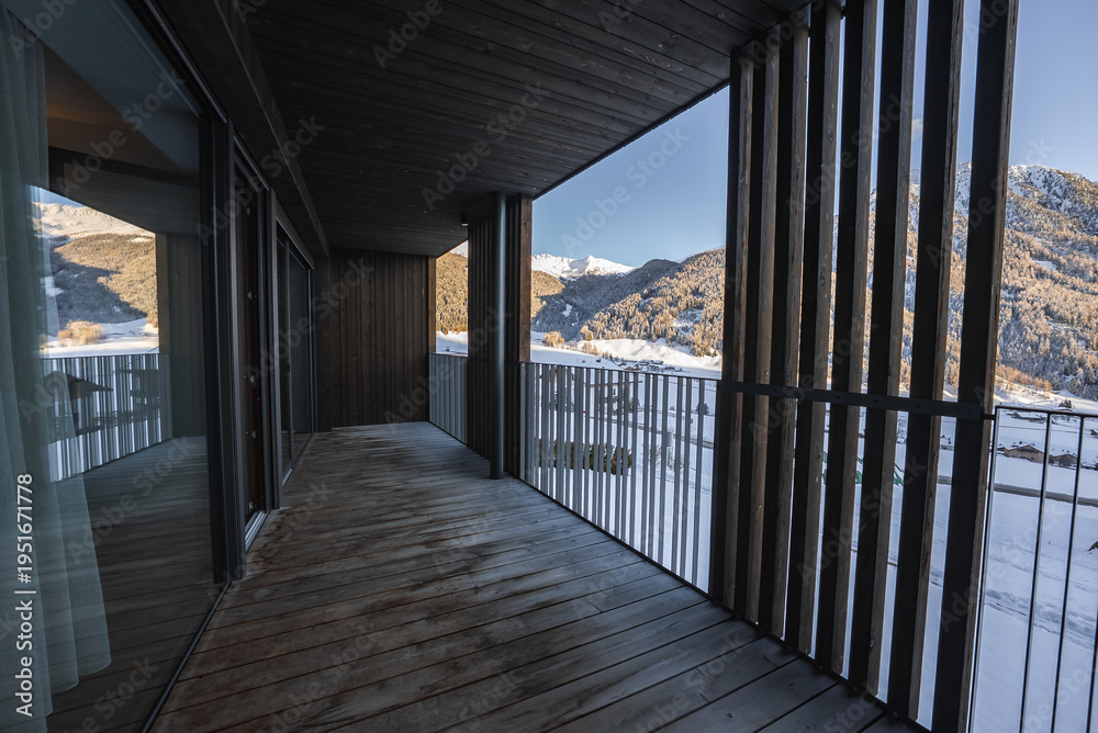 Obraz premium A spacious wooden balcony with vertical slatted screens faces the Dolomites in northern Italy as late afternoon light casts long shadows on the deck in winter.