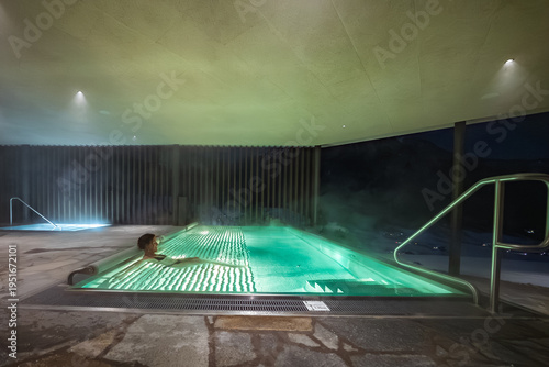 A person relaxes in a steaming illuminated outdoor spa pool under a modern canopy at a mountain hotel in the Dolomites, Italy, with peaks, mist, and soft low lighting.