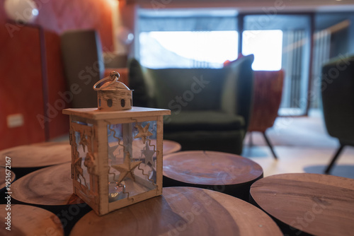 A small wooden lantern with star cutouts sits on a slice table in a modern Dolomites hotel lounge, soft warm lighting, blurred armchairs, snowy mountains through windows.