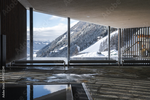 Minimalist daybeds line floor to ceiling glass as a hot pool reflects mountain light, metal handrails indicate access, steep forested slopes and groomed pistes define the Dolomites.
