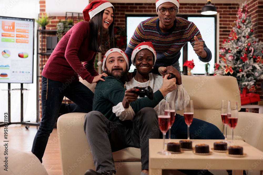 Obraz premium Happy coworkers having fun with video games at event in festive office decorated with xmas tree and ornaments. Playing competition on console and celebrating christmas holiday festivity.