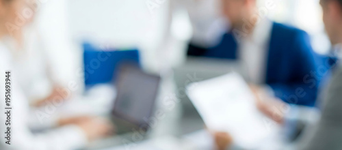 Blurred image of a business meeting with people discussing around a table, using laptops and documents, highlighting teamwork and collaboration.