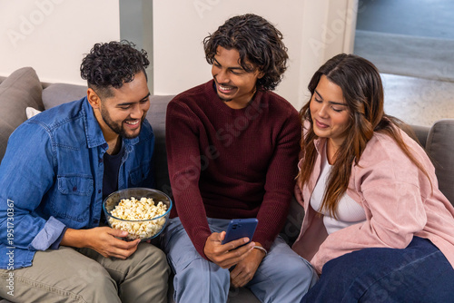 Three friends leaning in sharing smartphone and glass popcorn bowl on grey sofa in living room