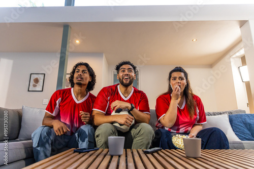 Diverse friends sitting on sofa in living room wearing red jerseys, holding soccer ball and popcorn