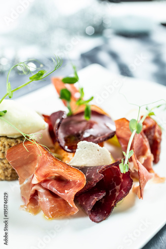 Modernly arranged appetizer with thinly sliced ​​prosciutto, cheese and side dishes, decorated with fresh sprouts on a white plate.