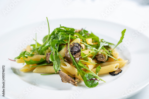 A portion of penne pasta served with black truffle leaves and fresh rocket on a white plate.