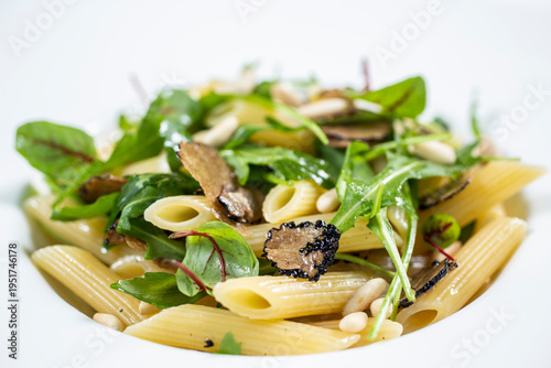A portion of penne pasta served with black truffle leaves and fresh rocket on a white plate.