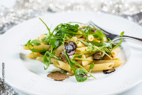 A portion of penne pasta served with black truffle leaves and fresh rocket on a white plate.