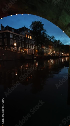 POV vertical video of a boat ride through an Amsterdam canal at twilight, passing under an illuminated historic brick bridge towards traditional Dutch grachten houses with glowing windows and street 