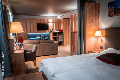 Modern lodge room at San Domenico in the Italian Alps presents wood paneling, plush double bed, blue sofa to a wall TV, soft lamps, desk, and apres ski mood.