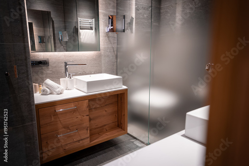 Warm wooden vanity, vessel sink, and chrome faucet sit by gray stone tiles. Frosted glass shower and soft indirect lighting evoke apres ski relaxation in winter.
